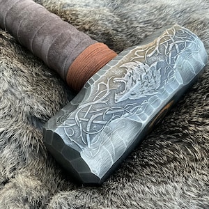 May include: A silver-colored hammer with a brown leather handle. The hammer head has a detailed design of a wolf's head and intricate knotwork.