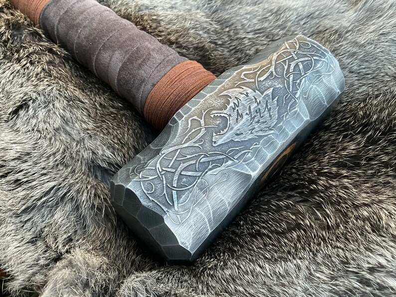 May include: A silver-colored hammer with a brown leather handle. The hammer head has a detailed design of a wolf's head and intricate knotwork.