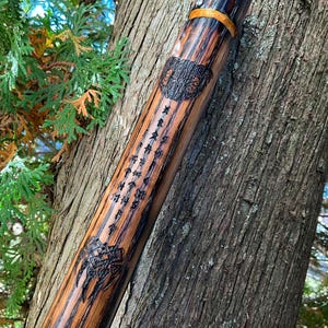 Op de afbeelding: Een houten wandelstok met een donkerbruine afwerking en ingewikkelde houtsnijwerk. De stok is versierd met een gouden band en runen.