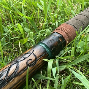 Puede incluir: Un bast&oacute;n de madera con intrincadas tallas y un acabado oscuro. El mango est&aacute; envuelto en cuero y gamuza marr&oacute;n, con una banda verde cerca de la parte superior. El bast&oacute;n est&aacute; sobre un fondo de hierba verde.