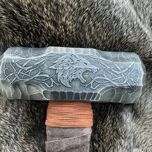 May include: A black metal hammer with a wolf head design and intricate carvings. The hammer has a brown leather wrap around the handle.