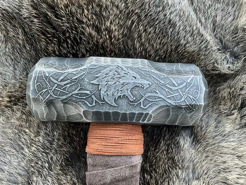 May include: A black metal hammer with a wolf head design and intricate carvings. The hammer has a brown leather wrap around the handle.