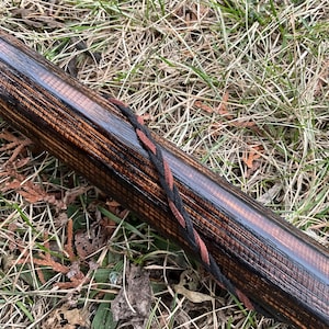 May include: A close-up of a wooden bow stave with a black and red braided cord wrapped around it. The wood has a dark brown stain and a natural grain pattern.