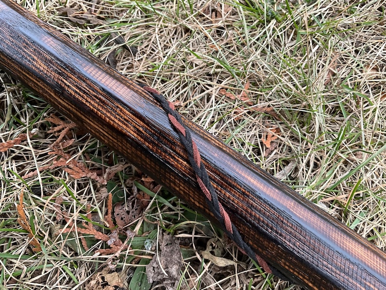 May include: A close-up of a wooden bow stave with a black and red braided cord wrapped around it. The wood has a dark brown stain and a natural grain pattern.