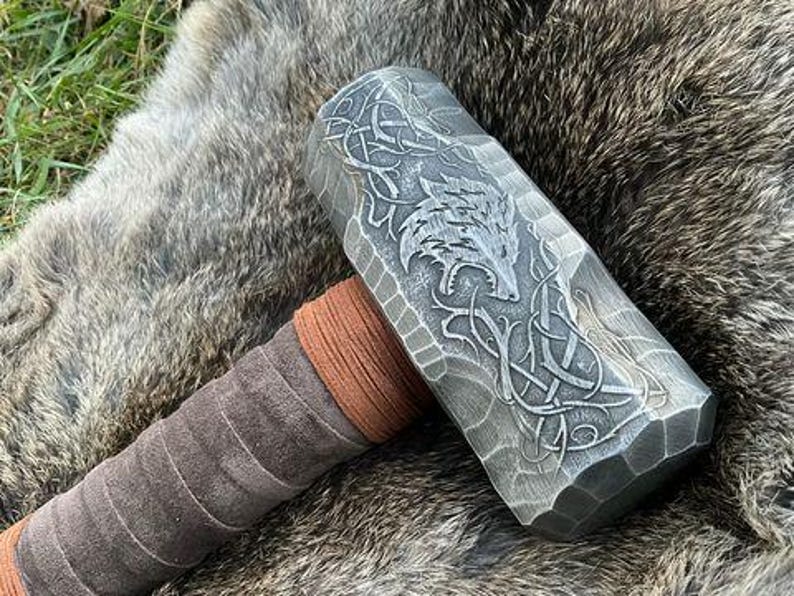 May include: A silver metal hammer with a brown leather handle. The hammer head has a detailed design of a wolf and a Celtic knot.