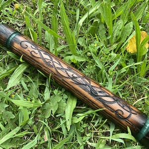 Puede incluir: Un bast&oacute;n de madera con intrincadas tallas de nudos celtas. El bast&oacute;n es de color marr&oacute;n oscuro con detalles negros y acentos verdes. Descansa sobre un lecho de hierba verde. El bast&oacute;n mide unos 60 cm.
