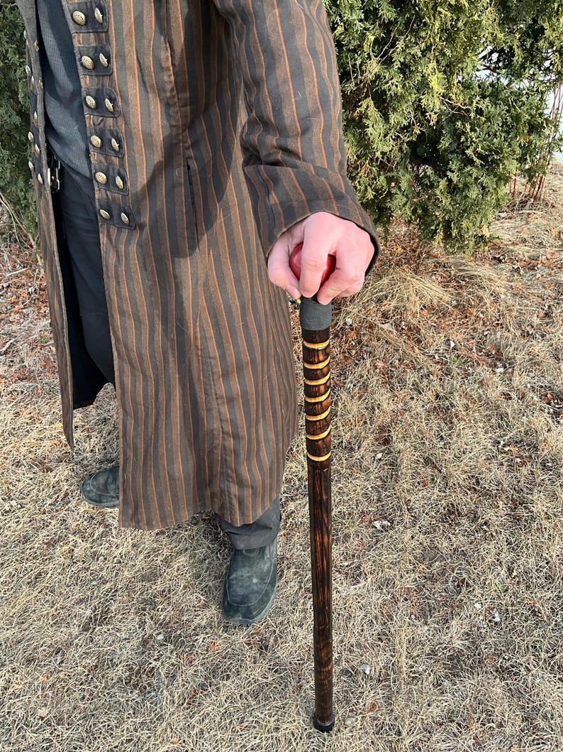 May include: A wooden walking stick with a black handle and gold accents. The stick is held by a person wearing a striped coat.