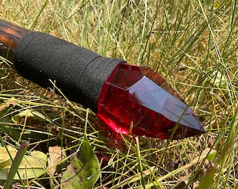 Puede incluir: Un bastón de madera con un agarre negro y una punta de cristal rojo grande y facetado.