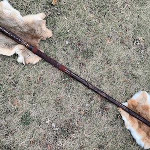 May include: A long, dark brown wooden staff with a gold band at the top and a black tip at the bottom. The staff is lying on a white and brown fur pelt.