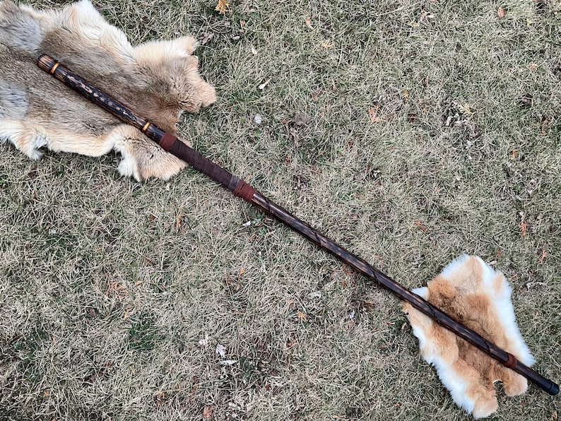 May include: A long, dark brown wooden staff with a gold band at the top and a black tip at the bottom. The staff is lying on a white and brown fur pelt.