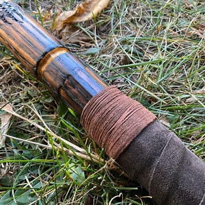 Op de afbeelding: Een houten staf met een donkerbruine vlek en een leren wikkel. De leren wikkel is bruin en heeft een ruwe textuur.