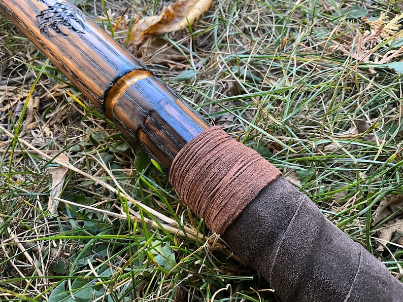 Op de afbeelding: Een houten staf met een donkerbruine vlek en een leren wikkel. De leren wikkel is bruin en heeft een ruwe textuur.