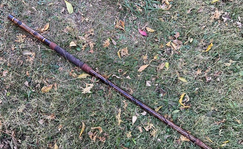 Op de afbeelding: Een lange, dunne houten staf met een donkerbruine vlek en zwarte en gouden accenten. De staf ligt op een bed van groen gras met bruine bladeren eromheen.