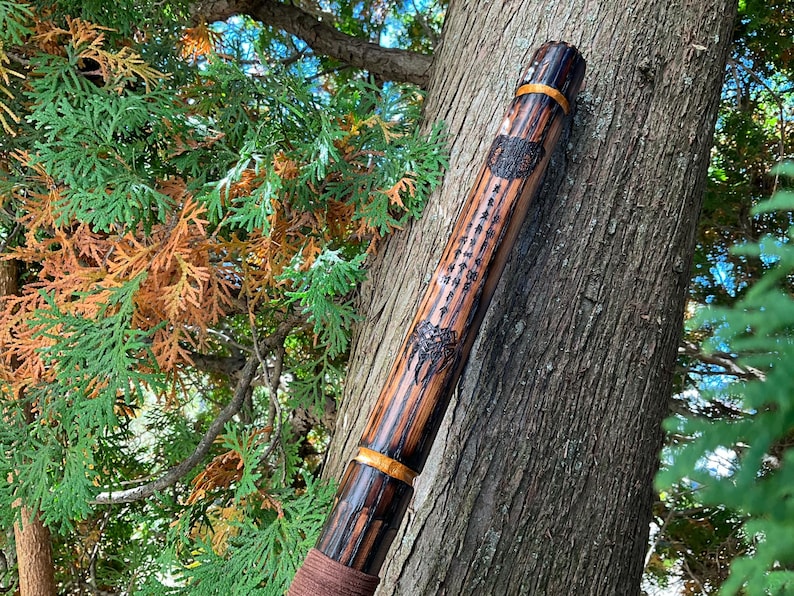 Op de afbeelding: Een houten wandelstok met een bruine leren wikkeling aan de onderkant en een gouden band dichtbij de bovenkant. De stok is versierd met een gesneden ontwerp en een zwart-witte afbeelding van een boom.