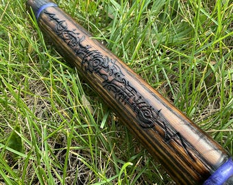 May include: A wooden wand with intricate, dark carvings along its length. The wand has a dark brown finish with a blue band near the handle. The wand is lying on a bed of green grass.