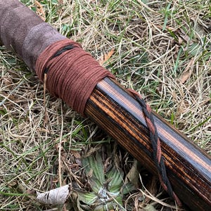 May include: Close-up of a wooden staff wrapped with brown and black leather cord. The staff has a dark brown stain and a natural wood grain pattern.