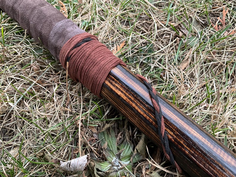 May include: Close-up of a wooden staff wrapped with brown and black leather cord. The staff has a dark brown stain and a natural wood grain pattern.
