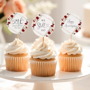 May include: Three cupcakes with white frosting on a white cake stand. Each cupcake has a decorative topper with floral accents and text that reads "50th Anniversary", "We Still Do", and "Happy Anniversary". A bouquet of flowers is in the background.