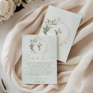 May include: Two white cards with gold text and a cross design, surrounded by a gold circle and green leaves. The cards are on a beige fabric with white flowers and greenery. The text on the card reads "Boda de Oro".