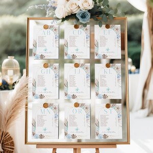 May include: A gold-framed seating chart displayed on a wooden easel. The chart features nine individual cards with guest names, adorned with floral and seashell designs. A bouquet of white and blue flowers tops the frame.