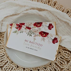 May include: Two white chocolate bar wrappers with gold edges, adorned with red floral arrangements and the text "MI BAUTIZO Lucia Fernandez 24 de Abril, 202x". The bars rest on a gray plate, atop a woven placemat.