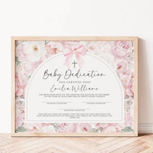 Pode incluir: Certificado de dedicação de bebê emoldurado com uma moldura de madeira clara. O certificado apresenta um design floral com flores rosa e um laço rosa. O texto inclui "Baby Dedication" e o nome "Emilia Williams".