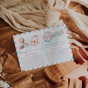 Könnte beinhalten: Eine druckbare Rezeptkarte mit blauem und rosa Rand. Die Karte zeigt eine Aquarell-Illustration eines Koffers, einer Karte, eines Hutes und einer Brille. Die Karte trägt die Wörter "Rezept", "Zutaten" und "Anleitung" aufgedruckt.