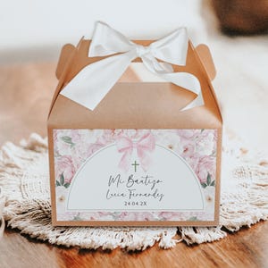 May include: A brown gift box with a white satin bow on top. The front of the box features a label with floral designs, a pink bow, a cross, and the text "Mi Bautizo Lucia Fernandez 24.04.2X."