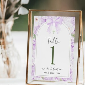 May include: A table number card in a gold frame. The card features a floral design with a lavender bow and cross. The text reads "Table 1" and "Emilia's Baptism, April 24th, 202X". The background is blurred, suggesting an event setting.