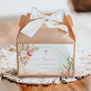 May include: A brown gift box tied with a white satin bow. The box features a label with floral and cactus illustrations, the text "mi Bautizo", a cross, and the name Lucia Fernandez with a date.