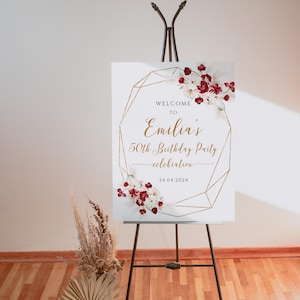 May include: A white welcome sign on a black easel, with gold geometric lines and floral accents. The sign reads "Welcome to Emilia's 50th Birthday Party celebration" with the date 24.04.202X. Dried flowers and a vase are in the foreground.