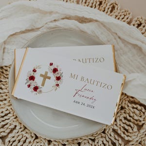Può includere: Due incarti rettangolari per barrette di cioccolato con sfondo bianco e bordi in lamina d'oro. Il design presenta una croce dorata circondata da illustrazioni floreali rosse e bianche. Il testo riporta MI BAUTIZO, Lucia Fernandez e ABR 24, 202X.