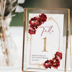 May include: A table number card in a gold frame. The card features a floral design with burgundy and pink flowers. The text on the card reads "Table 1" in gold, with additional text below indicating an anniversary date.