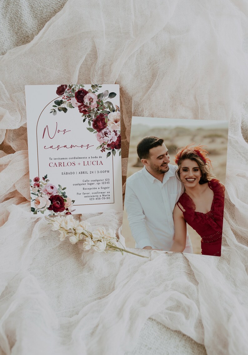 May include: A wedding invitation with a floral design in shades of burgundy, pink, and green. The invitation features a gold arch and the text "Nos casamos" in a cursive font. The text below the arch reads "Te invitamos cordialmente a boda de CARLOS + LUCIA SABADO ABRIL 24 13:00PM Calle cualquier 123 cualquier lugar, ep: 12345 Recepci&oacute;n a Seguir Por favor, confirmar su asistencia a Maria 123-456-78-90".