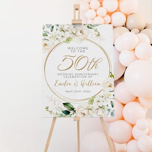 May include: A wooden easel displays a welcome sign for a 50th wedding anniversary. The sign features floral accents, gold lettering, and the names "Emilia & William." Peach and white balloons decorate the background.