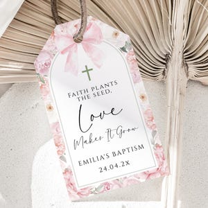 May include: A white tag with pink floral border and a cross. The text reads "Faith plants the seed, Love makes it grow. Emilia's Baptism 24.04.2X"