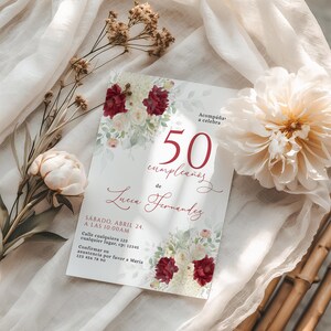 Puede incluir: Una tarjeta de invitación blanca con detalles florales y el texto "50 cumpleaños de Lucia Fernandez." La tarjeta presenta flores burdeos y blancas y está colocada sobre una tela suave y drapeada con elementos florales adicionales.