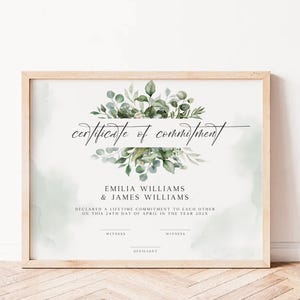 Può includere: Un certificato di impegno incorniciato in una cornice di legno chiaro. Il certificato presenta le parole "certificate of commitment" in elegante scrittura sopra i nomi Emilia Williams & James Williams. Il fogliame verde adorna la parte superiore.