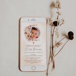Könnte beinhalten: Ein weißes Smartphone zeigt eine digitale Taufeinladung für Carlos Fernandez mit einem kreisförmigen Babyfoto, umrahmt von getrockneten Blumen, einem kleinen Kreuzsymbol und Veranstaltungsdetails in spanischer Sprache auf beigem Grund.