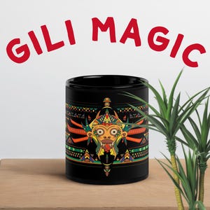 Könnte beinhalten: Eine schwarze Keramik-Tasse mit einem farbenfrohen Stammesdesign. Das Design zeigt ein zentrales Gesicht mit orangefarbenen, gelben und grünen Akzenten. Die Worte "GILI MAGIC" stehen in Rot über der Tasse. Die Tasse steht auf einer Holzoberfläche.