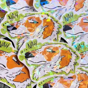 May include: A collection of stickers featuring a dog's head with orange and white fur, a black nose, and the word "NAH" in a green bubble. The stickers have a holographic effect and are arranged on a textured green surface.