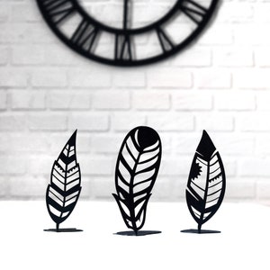 May include: Three black metal feather-shaped sculptures on a white surface. Each sculpture has a unique design and is about 6 inches tall.
