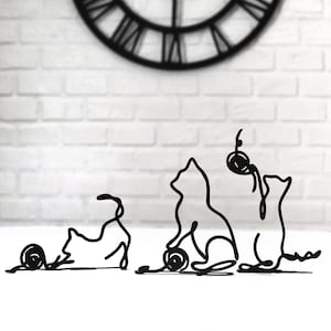 May include: Three black metal cat sculptures with a minimalist design. The cats are posed in different positions, one with a ball of yarn, one sitting and one lying down.