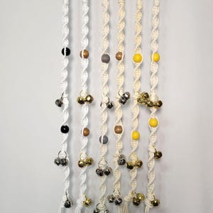 May include: Macrame wall hanging with multiple strands. Each strand features a spiral design, adorned with beads in black, brown, gray, and yellow, and small silver and gold bells. The strands are suspended from a wooden dowel.