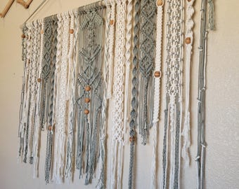 Puede incluir: Un tapiz de macramé en blanco y gris, con intrincados patrones de nudos y cuentas de madera. La obra de arte textil está suspendida de una barra de madera, creando un estilo bohemio. Cuelga sobre una pared de color neutro.