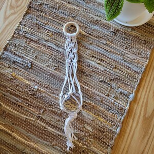 May include: White macrame plant hanger with a wooden ring at the top and a tassel at the bottom. The hanger is hanging on a woven rug.