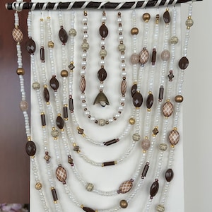 May include: A brown and white beaded wall hanging with a bell charm. The hanging features multiple strands of beads in various shapes and sizes, including round, oval, and rectangular beads.
