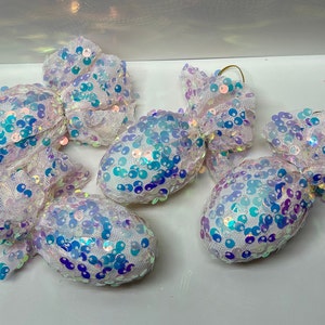 May include: Four white candy cane ornaments covered in iridescent sequins. The ornaments are tied with white tulle ribbon and have a gold wire loop for hanging.