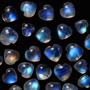Puede incluir: Una colecci&oacute;n de peque&ntilde;os cabujones de piedra lunar en forma de coraz&oacute;n. Cada piedra presenta una base transl&uacute;cida de color blanco hueso con una distintiva adularescencia azul brillante que var&iacute;a en intensidad sobre las superficies lisas, pulidas y redondeadas.