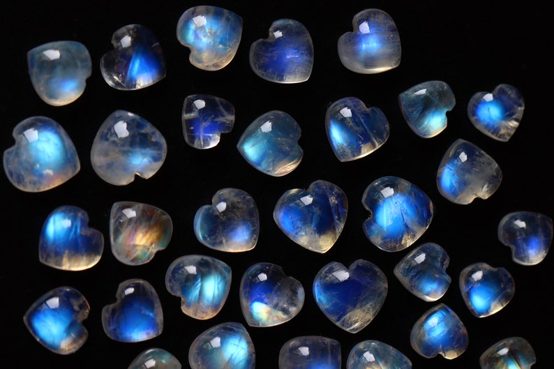 Puede incluir: Una colecci&oacute;n de peque&ntilde;os cabujones de piedra lunar en forma de coraz&oacute;n. Cada piedra presenta una base transl&uacute;cida de color blanco hueso con una distintiva adularescencia azul brillante que var&iacute;a en intensidad sobre las superficies lisas, pulidas y redondeadas.